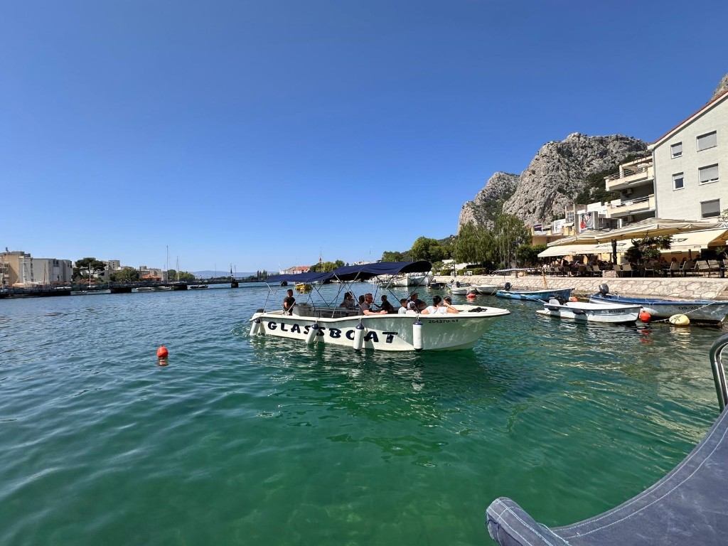 Glass boat with passengers on turquoise water of the Cetina River in Omiš, waterfront and cliffs behind