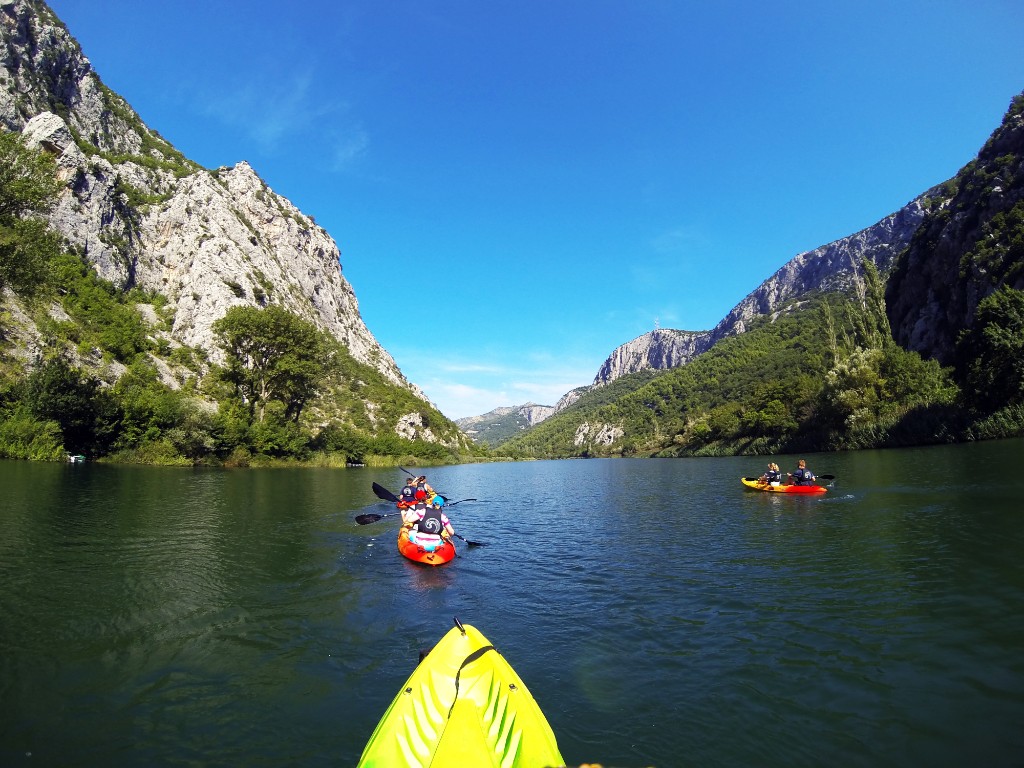 Kayakers on the calm Cetina River between tall limestone canyon cliffs, first-person view from a yellow kayak bow